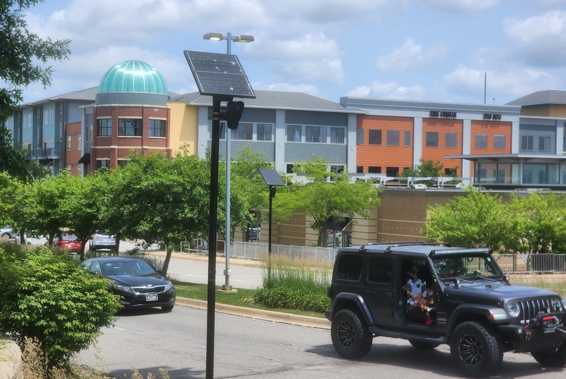 A two-way divided secondary road accessing a commercial are shown. In the foreground, which is a curbside, a green, leafy bush is shown on the bottom left, and a black pole topped with solar panel and Flock ALPR camera installed. In the midground, two vehicles can be seen driving right, a dark grey Jeep in front, a black Kia sedan behind. Behind the road and cars is a median featuring long, decorative grasses on the right side, medium-size trees on the left side. In the middle of the median, near a two-headed street lamp, another black pole topped with solar panel and Flock ALPR camera can be seen. The background shows multi-color, multi-story commercial buildings, the corner of which features a large, teal blue dome of semi-transparent material.