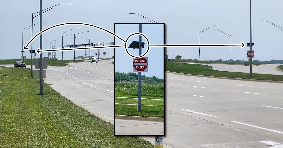 Roadside depicted, left foreground shows green grass along the curbside. Lots of street lamp and traffic signal poles are shown. In the center, an enhanced example of the median installed Flock ALPR system is inserted as a graphic overlay. Black arrows point out, one to the right, two to the left, identifying the ALPR systems installed in the road area.