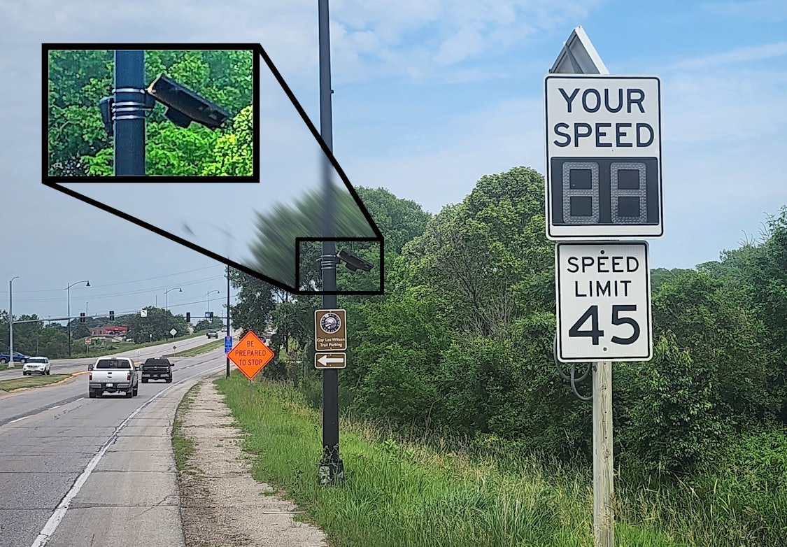 The side of a roadway is shown, in the foreground is an electronic vehicle speed notification sign along with a speed limit sign. Immediately behind is a street lamp pole with an attached Flock Falcon™ ALPR camera system, solar-powered.